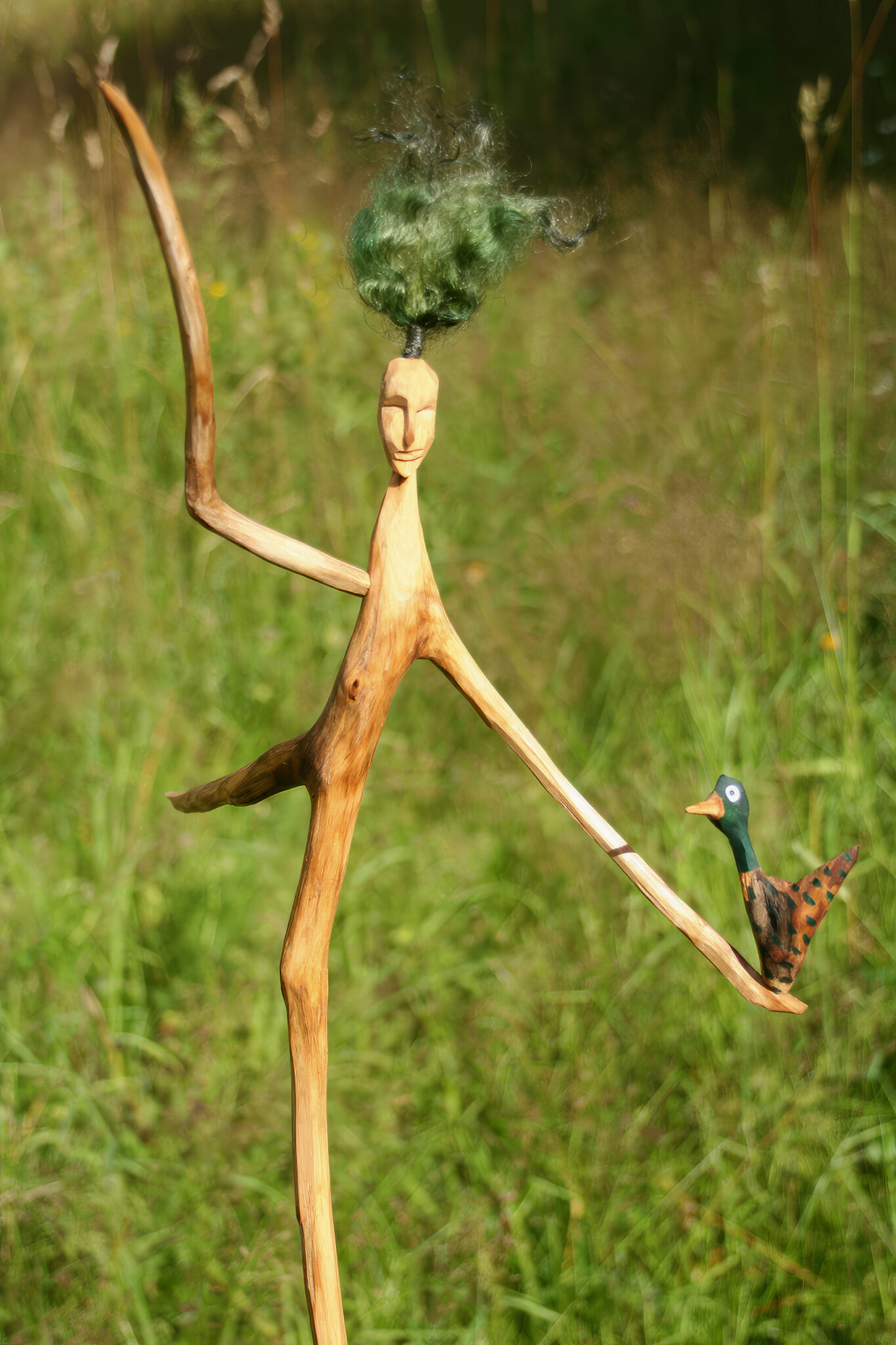 En pinnskulptur i en äng, håller en gräsand i ena handen.Foto.